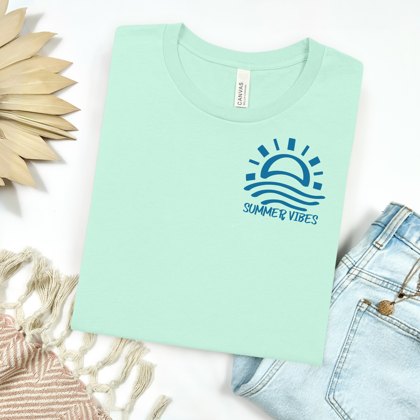 Bella and Canvas 3001 "Summer Vibes" t shirt with sun and waves one color graphic. Picture on a flay lay mockup with pair of blue jeans and blanket and a wicker sun shaped accessory.