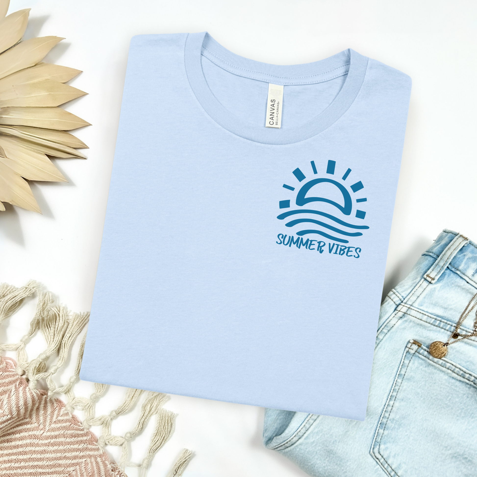 Bella and Canvas 3001 "Summer Vibes" t shirt with sun and waves one color graphic. Picture on a flay lay mockup with pair of blue jeans and blanket and a wicker sun shaped accessory.