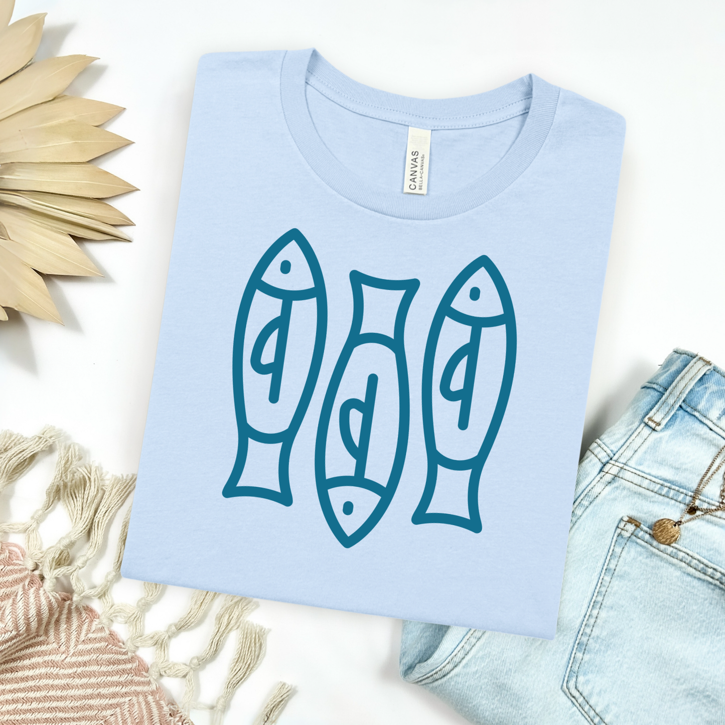 Bella canvas t shirt of a monoline art of  three sardines in one color graphic. Picture on a flay lay mockup with pair of blue jeans and blanket and a wicker sun shaped accessories.