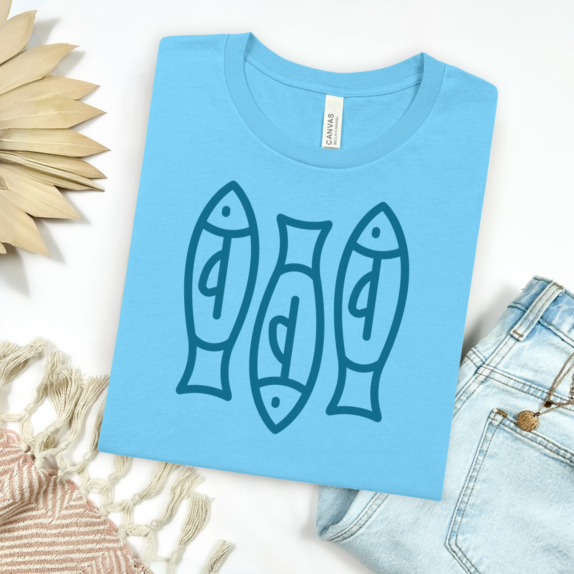 Bella canvas t shirt of a monoline art of  three sardines in one color graphic. Picture on a flay lay mockup with pair of blue jeans and blanket and a wicker sun shaped accessories.