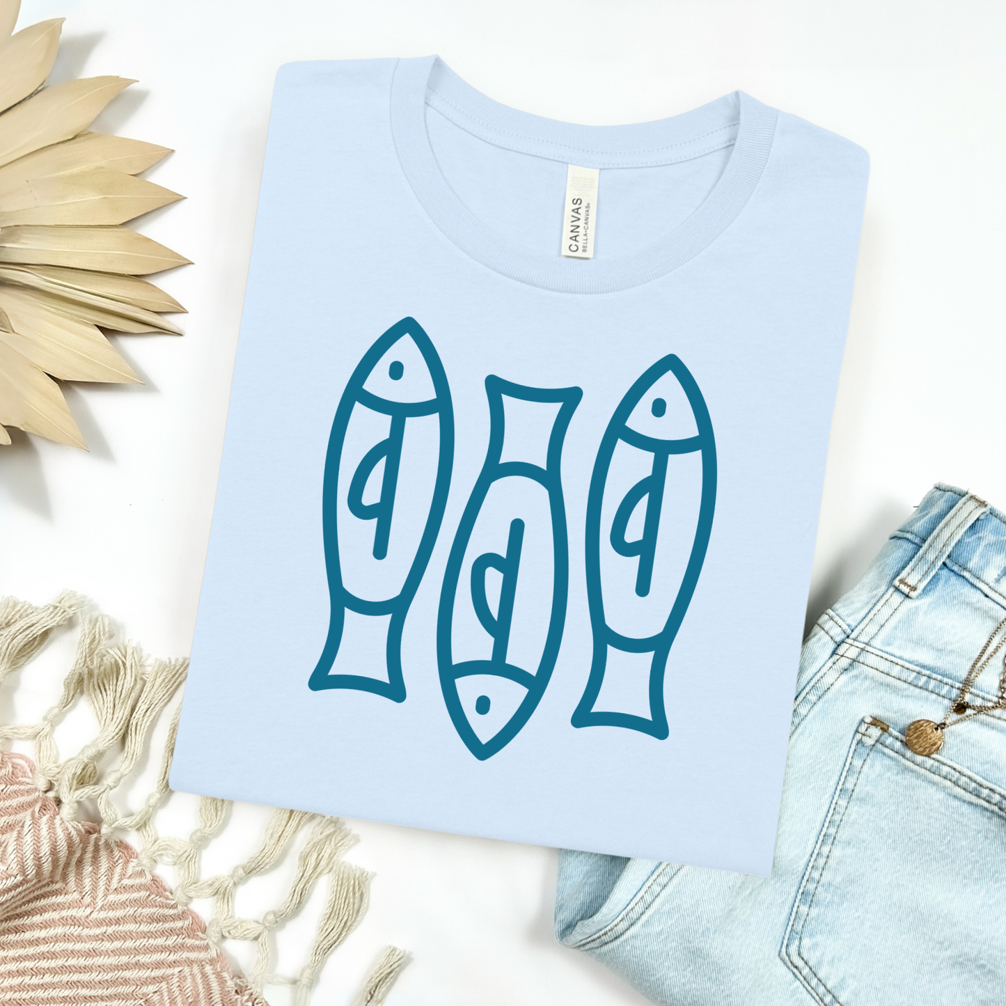 Bella canvas t shirt of a monoline art of  three sardines in one color graphic. Picture on a flay lay mockup with pair of blue jeans and blanket and a wicker sun shaped accessories.