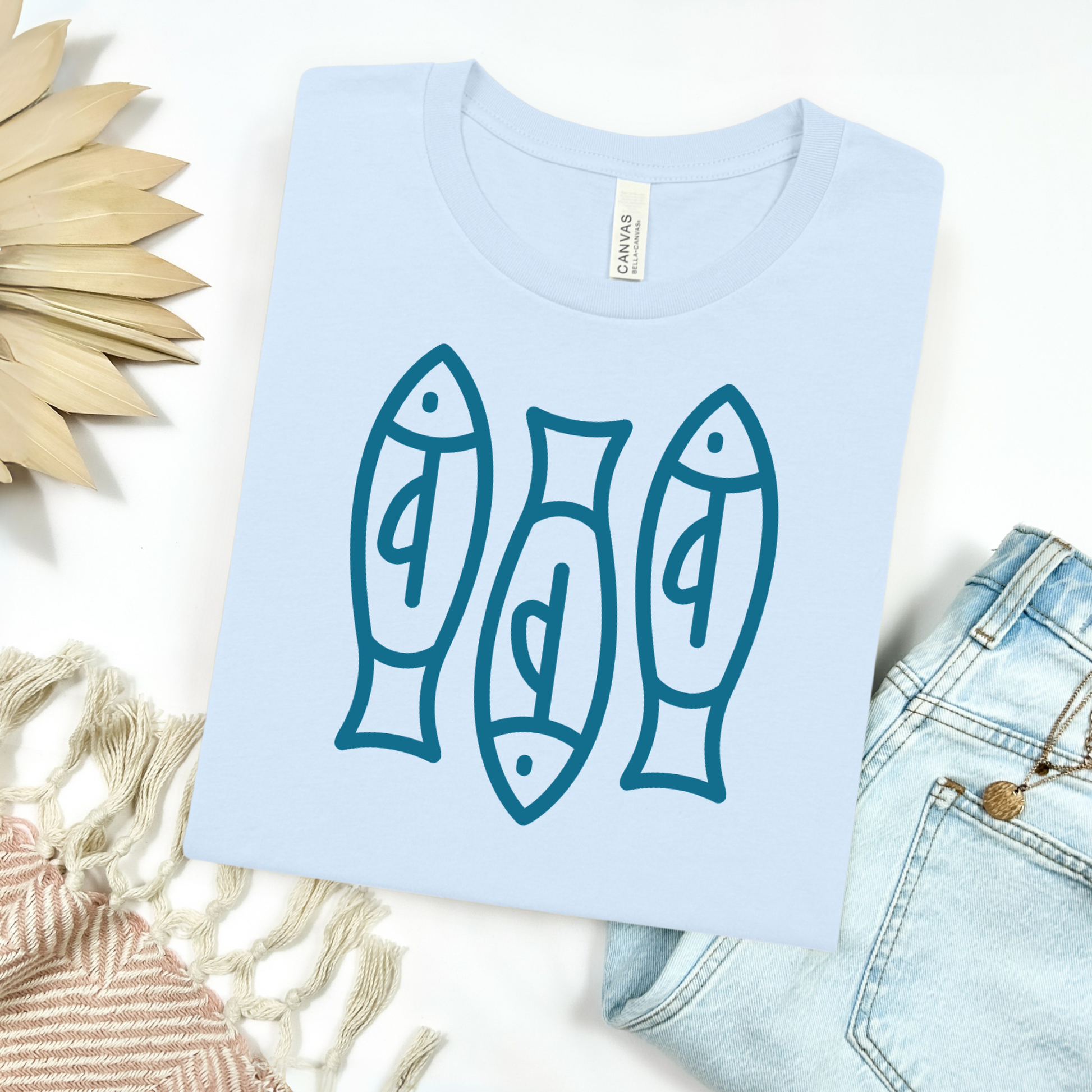 Bella canvas t shirt of a monoline art of  three sardines in one color graphic. Picture on a flay lay mockup with pair of blue jeans and blanket and a wicker sun shaped accessories.