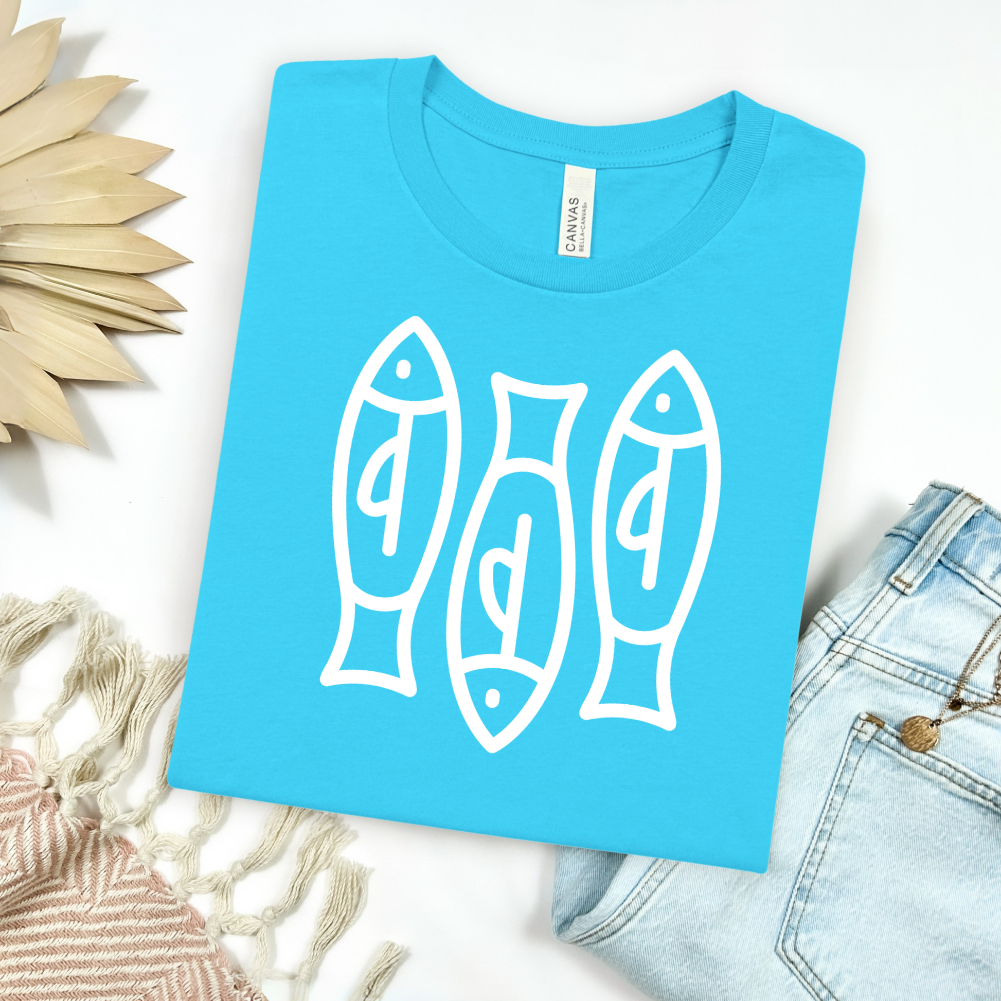 Bella canvas t shirt of a monoline art of  three sardines in one color graphic. Picture on a flay lay mockup with pair of blue jeans and blanket and a wicker sun shaped accessories.