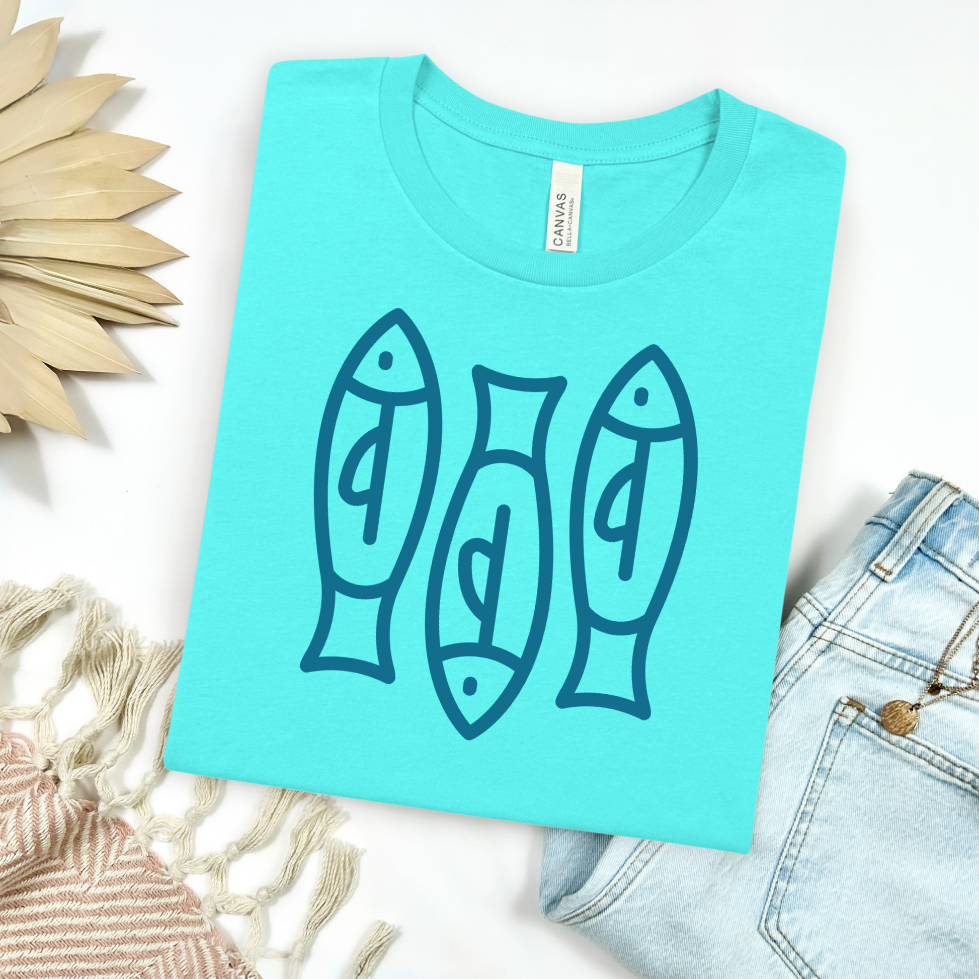 Bella canvas t shirt of a monoline art of  three sardines in one color graphic. Picture on a flay lay mockup with pair of blue jeans and blanket and a wicker sun shaped accessories.
