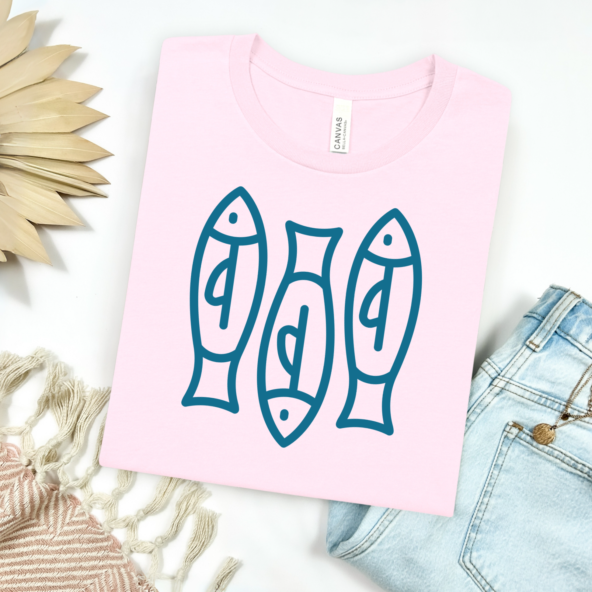 Bella canvas t shirt of a monoline art of  three sardines in one color graphic. Picture on a flay lay mockup with pair of blue jeans and blanket and a wicker sun shaped accessories.