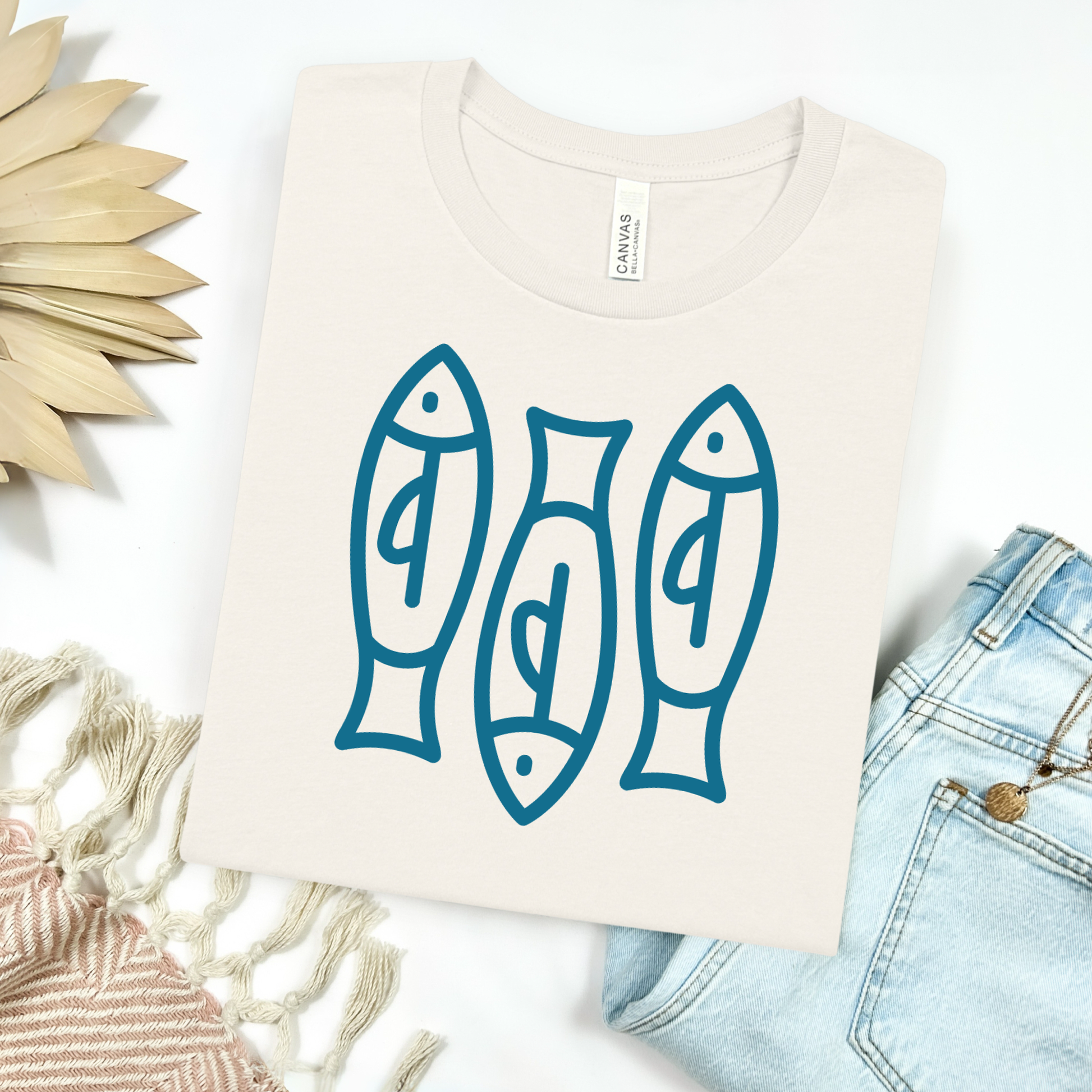 Bella canvas t shirt of a monoline art of  three sardines in one color graphic. Picture on a flay lay mockup with pair of blue jeans and blanket and a wicker sun shaped accessories.