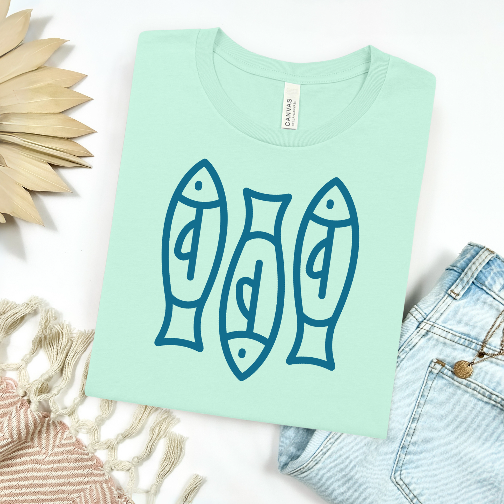 Bella canvas t shirt of a monoline art of  three sardines in one color graphic. Picture on a flay lay mockup with pair of blue jeans and blanket and a wicker sun shaped accessories.