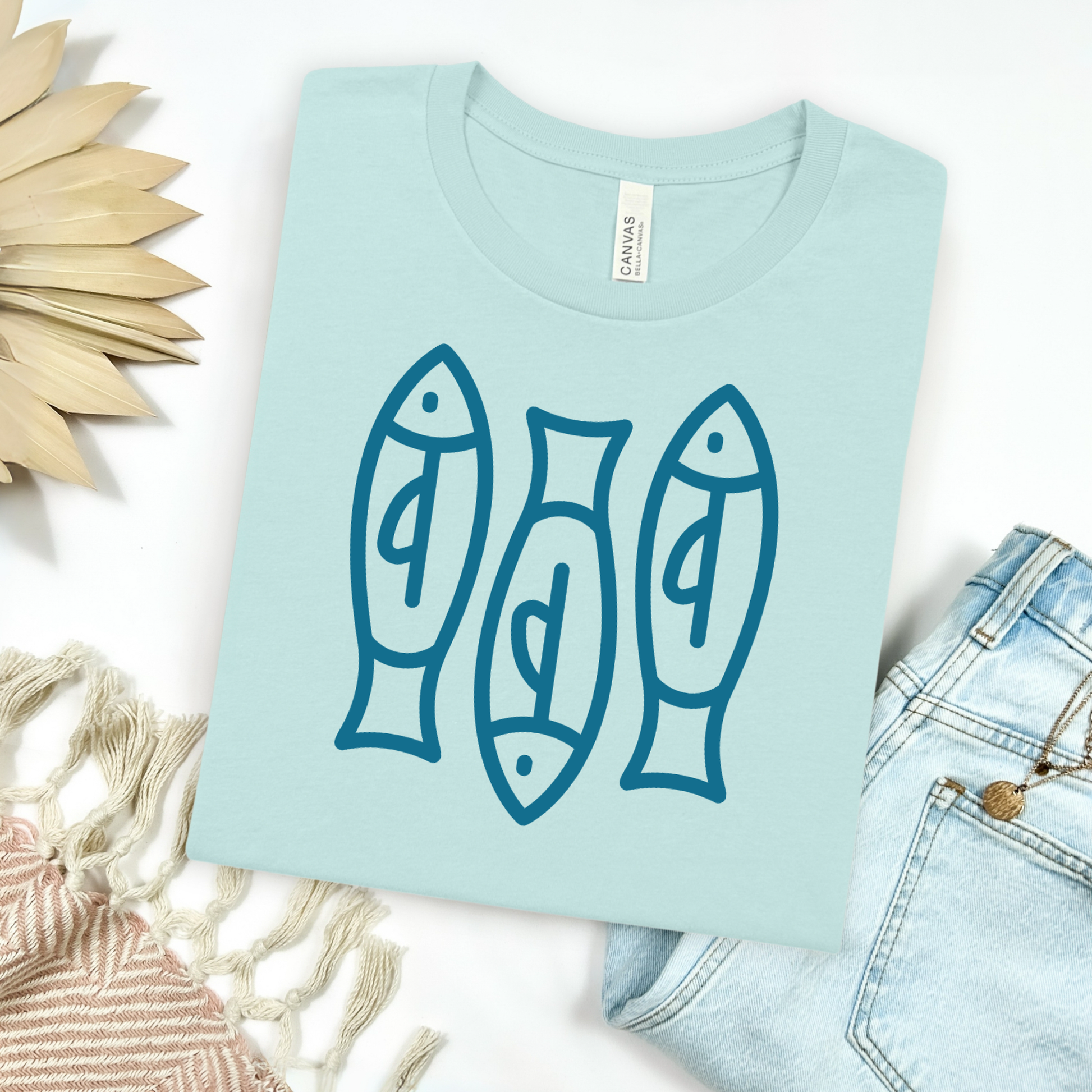Bella canvas t shirt of a monoline art of  three sardines in one color graphic. Picture on a flay lay mockup with pair of blue jeans and blanket and a wicker sun shaped accessories.