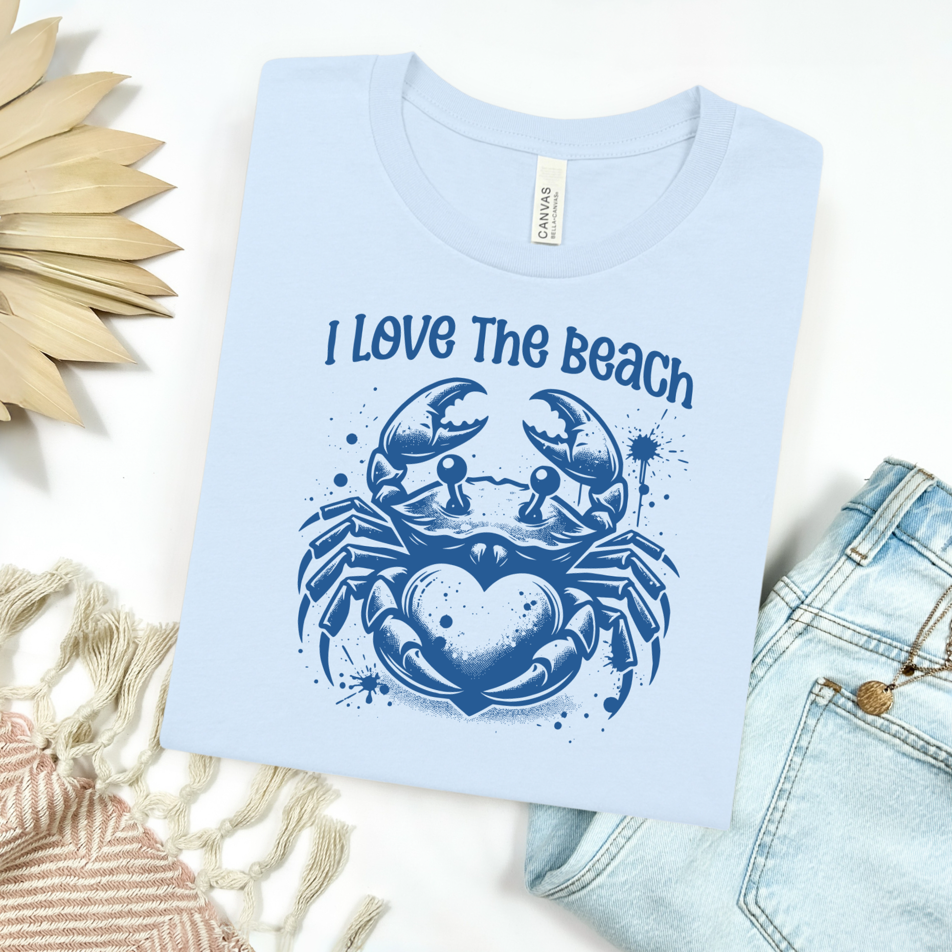 Bella Canvas 3001 "I Love The Beach" t shirt with a crab holding a heart one color graphic. Picture on a flay lay mockup with pair of blue jeans and blanket and a wicker sun shaped accessory.