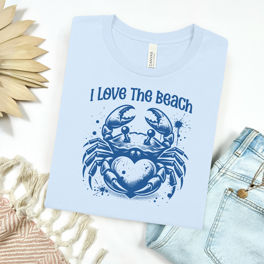 Bella Canvas 3001 "I Love The Beach" t shirt with a crab holding a heart one color graphic. Picture on a flay lay mockup with pair of blue jeans and blanket and a wicker sun shaped accessory.