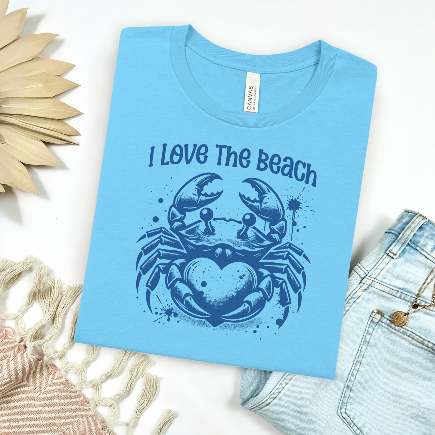 Bella Canvas 3001 "I Love The Beach" t shirt with a crab holding a heart one color graphic. Picture on a flay lay mockup with pair of blue jeans and blanket and a wicker sun shaped accessory.