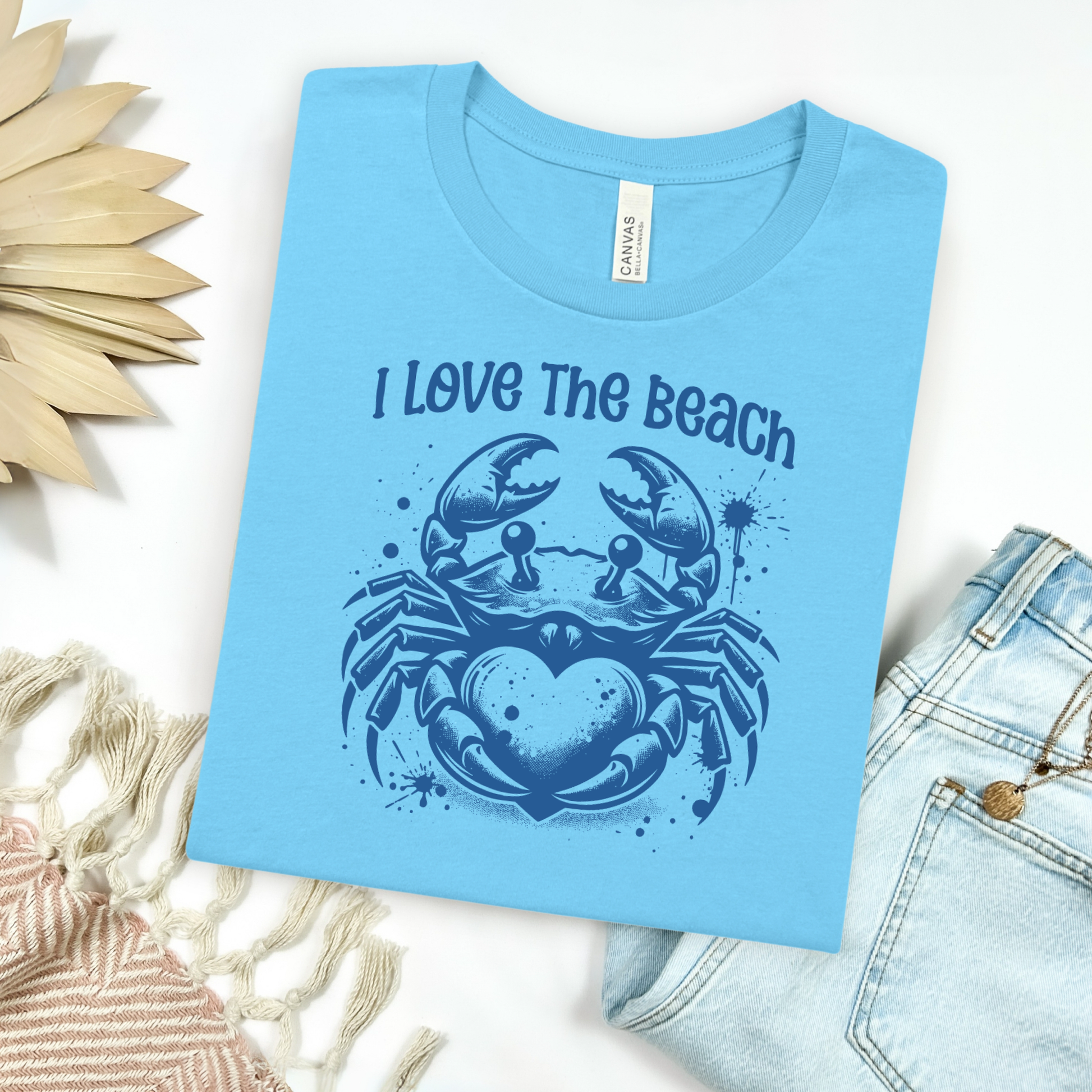 Bella Canvas 3001 "I Love The Beach" t shirt with a crab holding a heart one color graphic. Picture on a flay lay mockup with pair of blue jeans and blanket and a wicker sun shaped accessory.