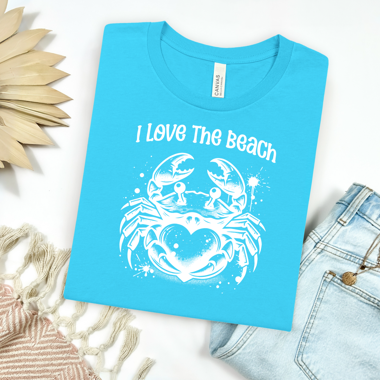 Bella Canvas 3001 "I Love The Beach" t shirt with a crab holding a heart one color graphic. Picture on a flay lay mockup with pair of blue jeans and blanket and a wicker sun shaped accessory.