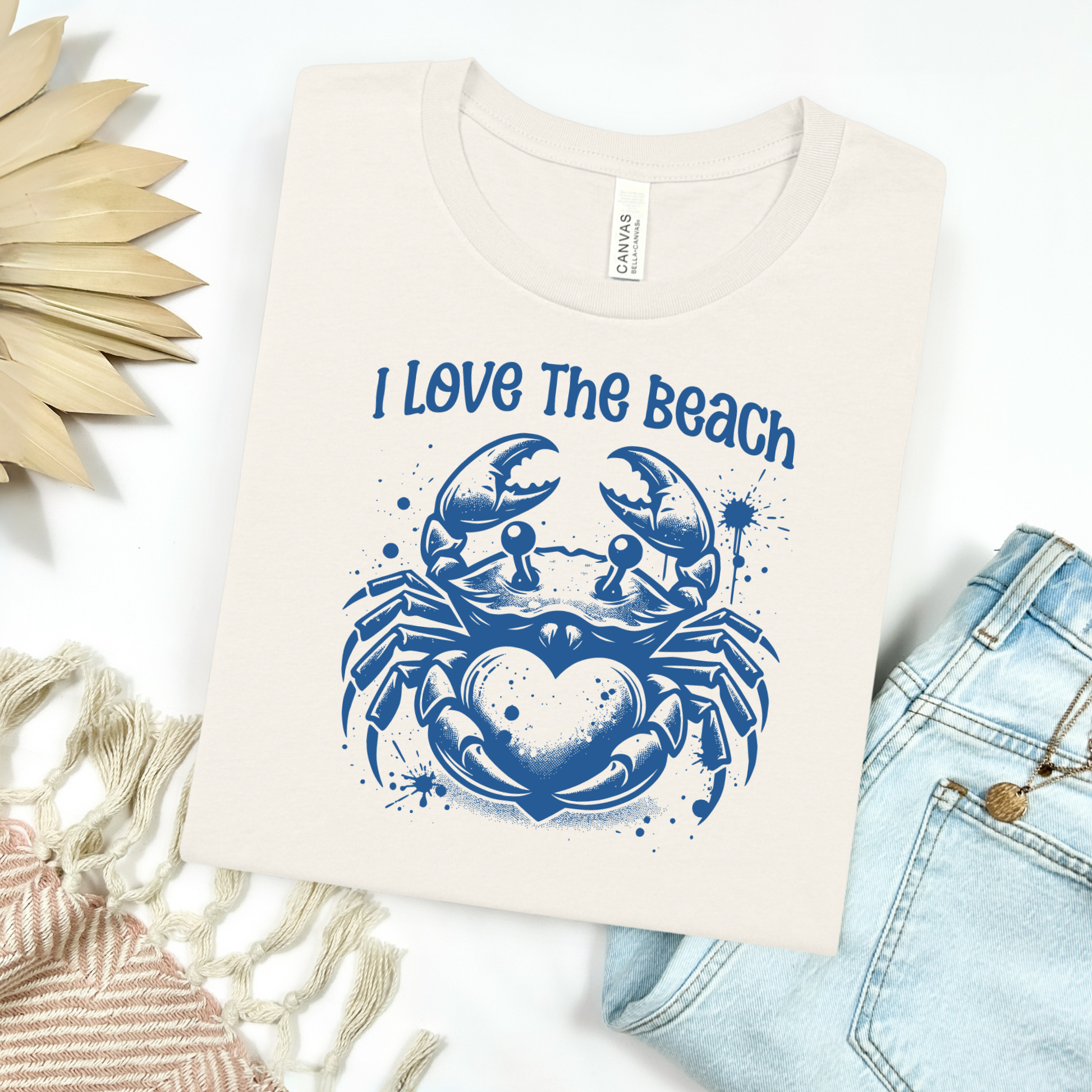 Bella Canvas 3001 "I Love The Beach" t shirt with a crab holding a heart one color graphic. Picture on a flay lay mockup with pair of blue jeans and blanket and a wicker sun shaped accessory.