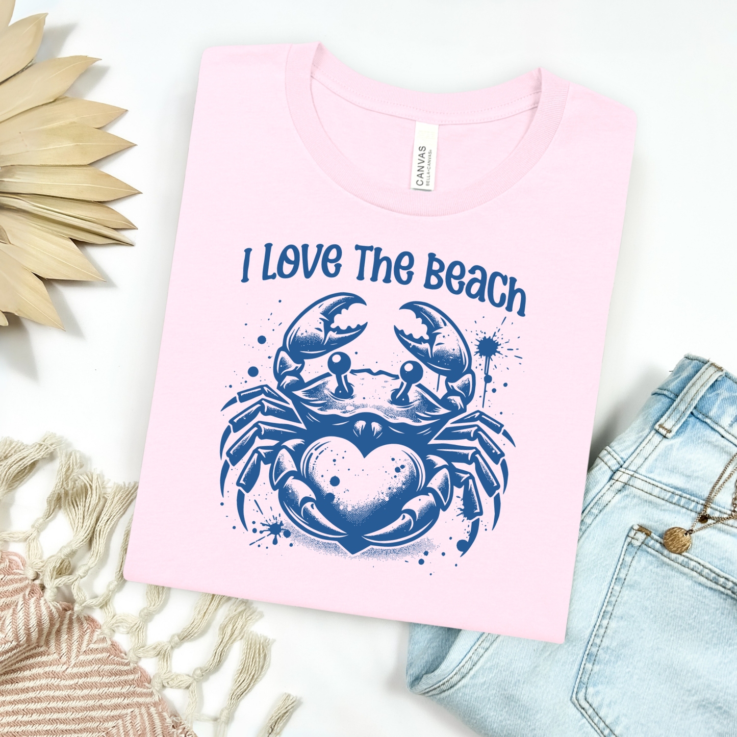 Bella Canvas 3001 "I Love The Beach" t shirt with a crab holding a heart one color graphic. Picture on a flay lay mockup with pair of blue jeans and blanket and a wicker sun shaped accessory.
