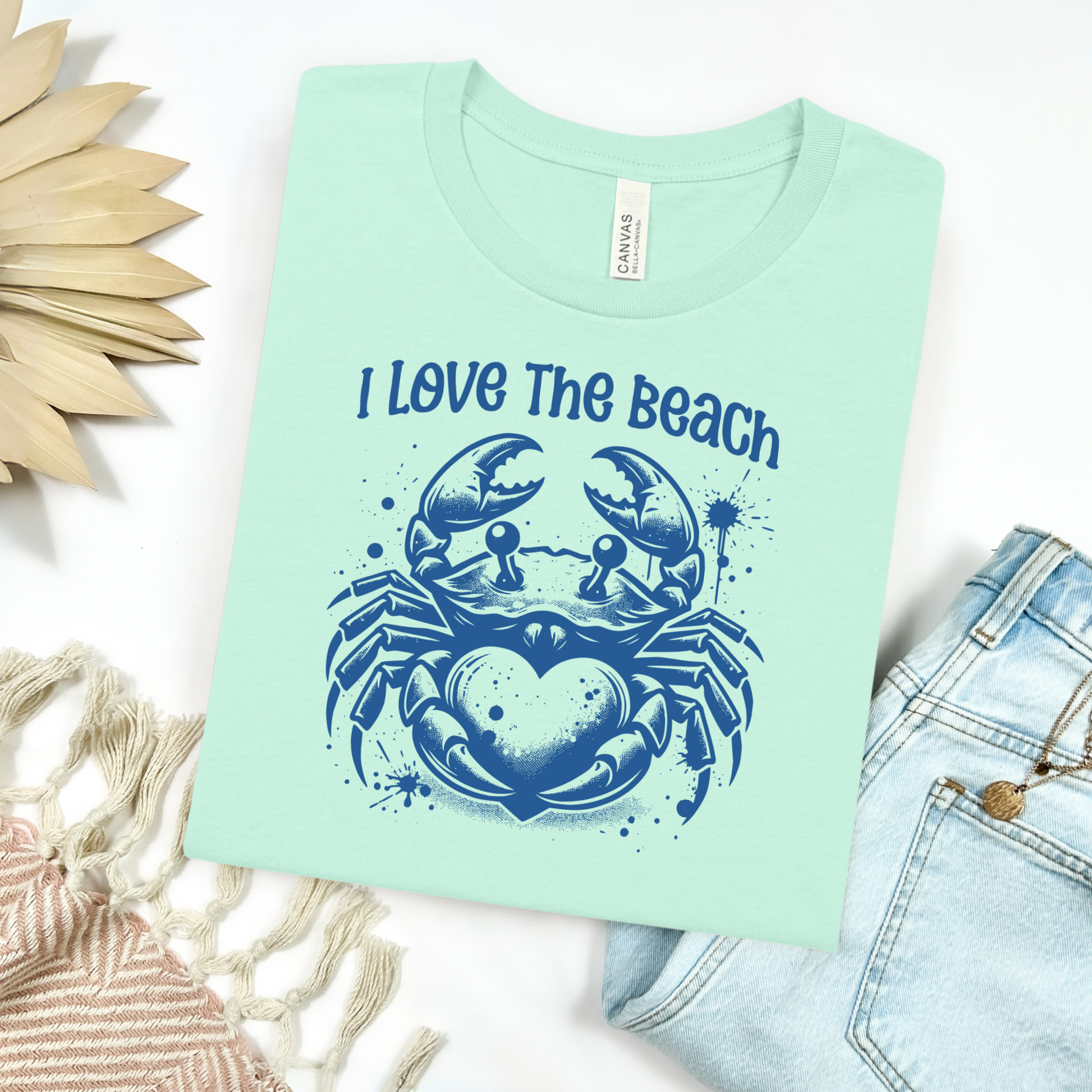 Bella Canvas 3001 "I Love The Beach" t shirt with a crab holding a heart one color graphic. Picture on a flay lay mockup with pair of blue jeans and blanket and a wicker sun shaped accessory.
