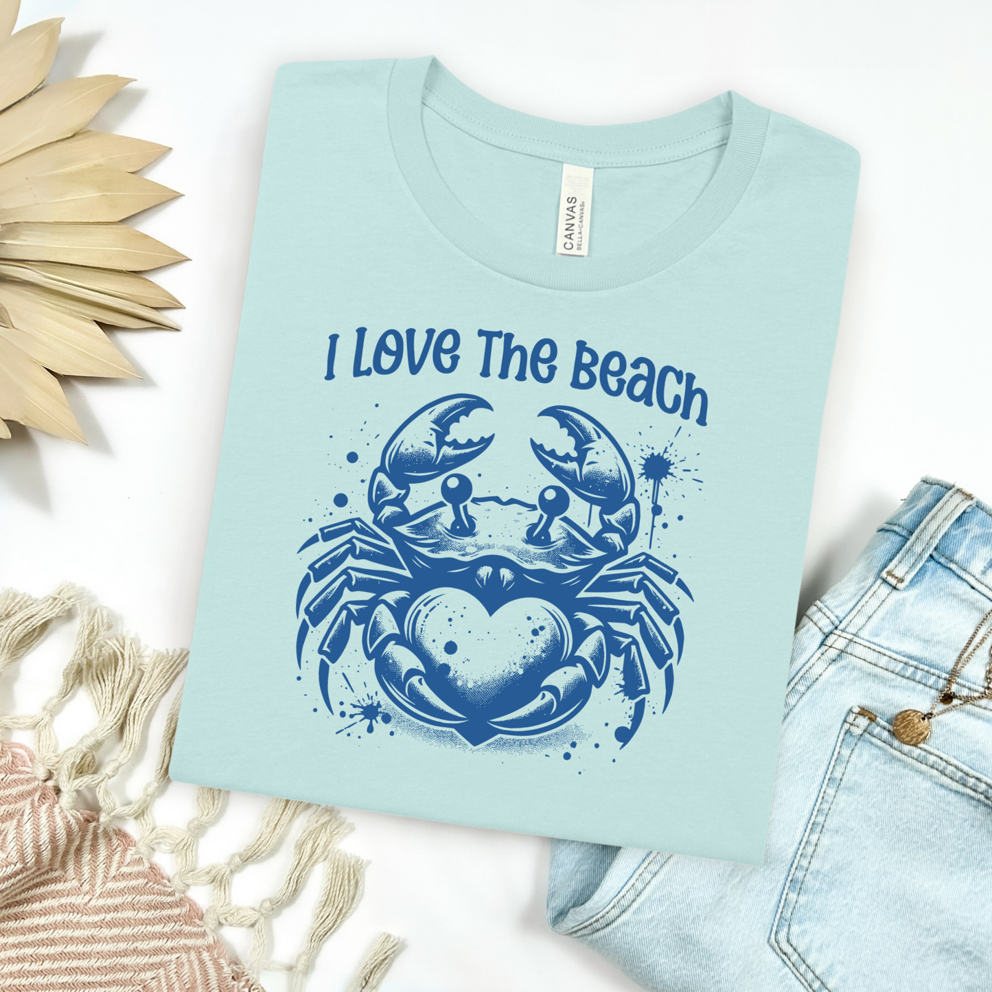 Bella Canvas 3001 "I Love The Beach" t shirt with a crab holding a heart one color graphic. Picture on a flay lay mockup with pair of blue jeans and blanket and a wicker sun shaped accessory.