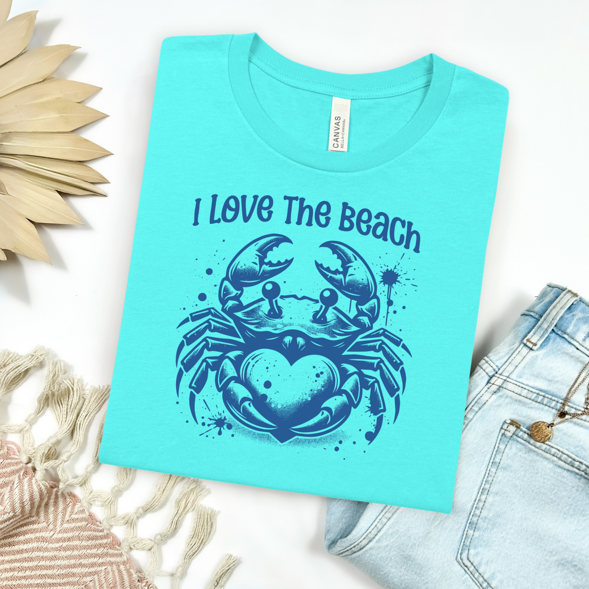 Bella Canvas 3001 "I Love The Beach" t shirt with a crab holding a heart one color graphic. Picture on a flay lay mockup with pair of blue jeans and blanket and a wicker sun shaped accessory.