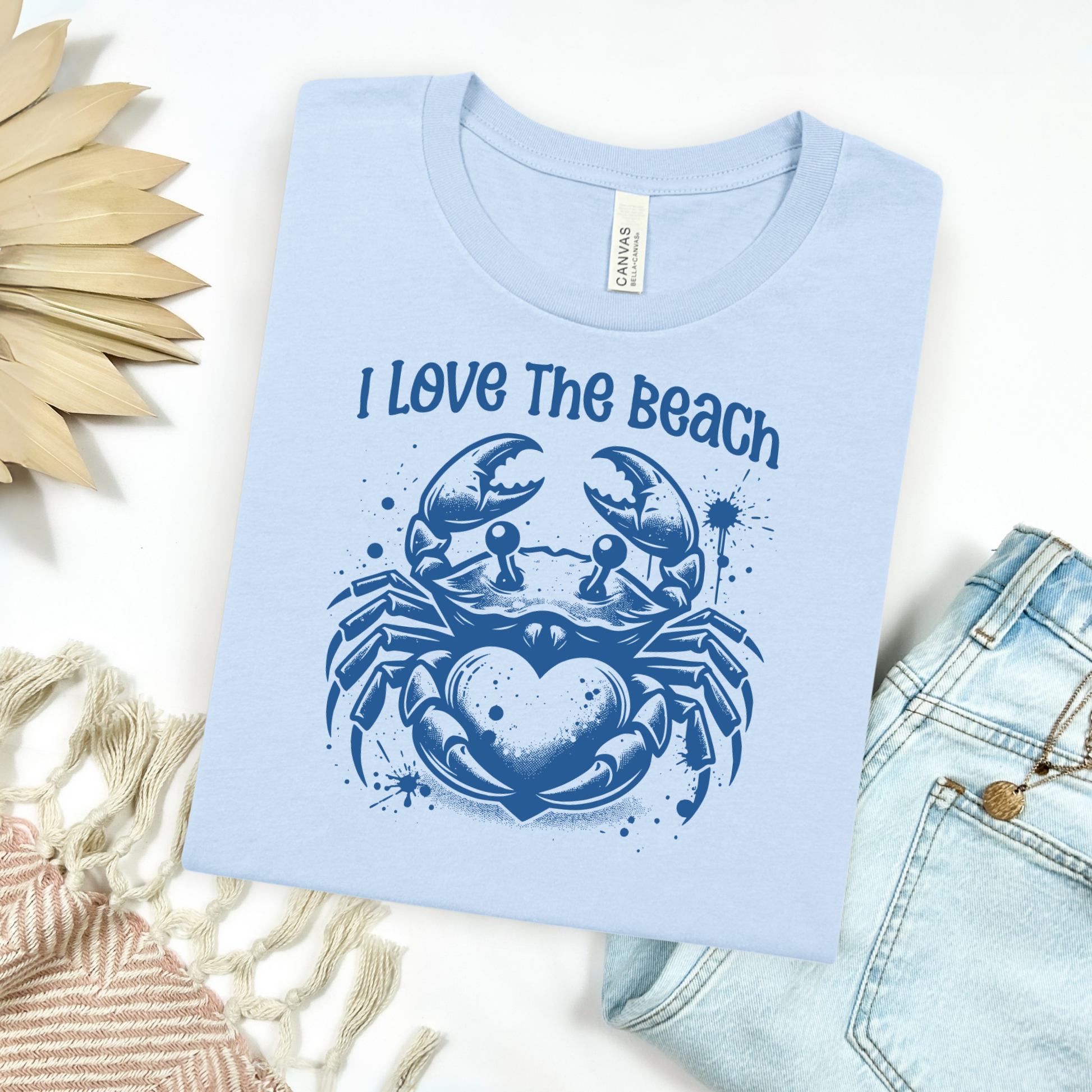 Bella Canvas 3001 "I Love The Beach" t shirt with a crab holding a heart one color graphic. Picture on a flay lay mockup with pair of blue jeans and blanket and a wicker sun shaped accessory.