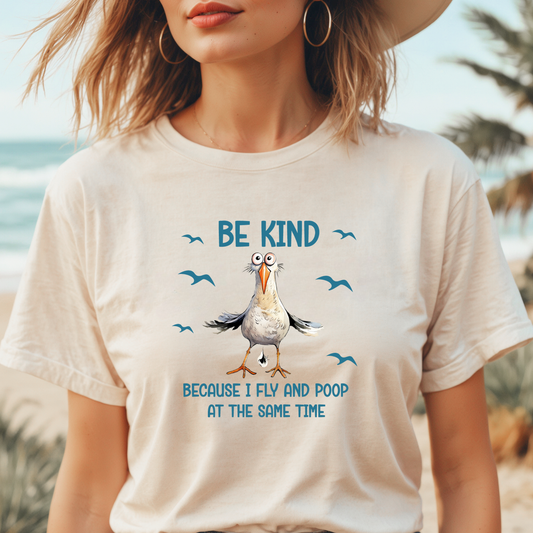 Person wearing a t-shirt with a cartoon seagull and text 'Be kind because I fly and poop at the same time' on a beach.