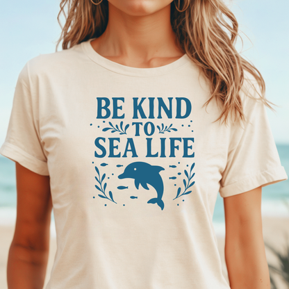 Person wearing a t-shirt with 'Be Kind to Sea Life' and a dolphin graphic on a beach.