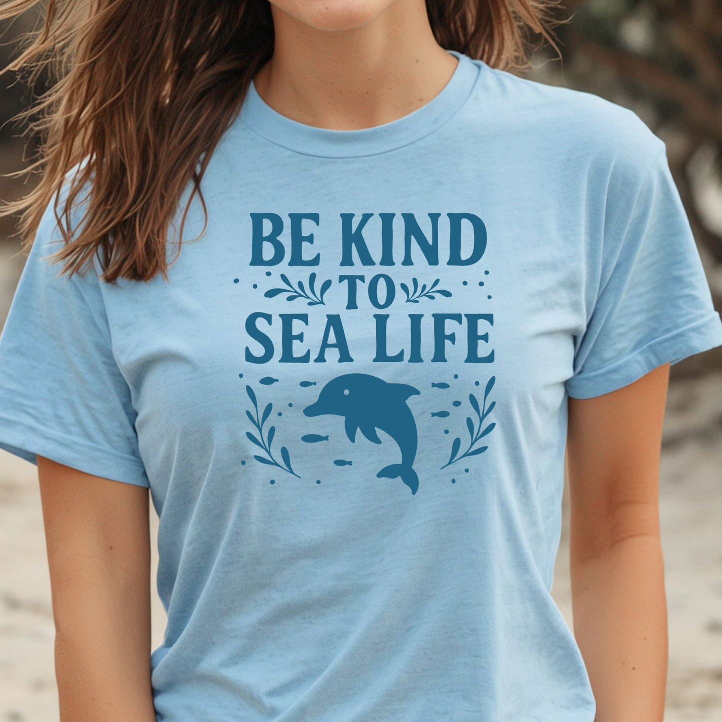 Light blue t-shirt with 'Be Kind to Sea Life' text and dolphin graphic on a beach.