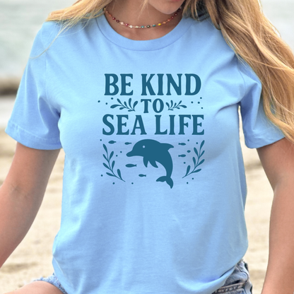 Person wearing a light blue t-shirt with 'Be Kind to Sea Life' and a dolphin graphic on a beach.