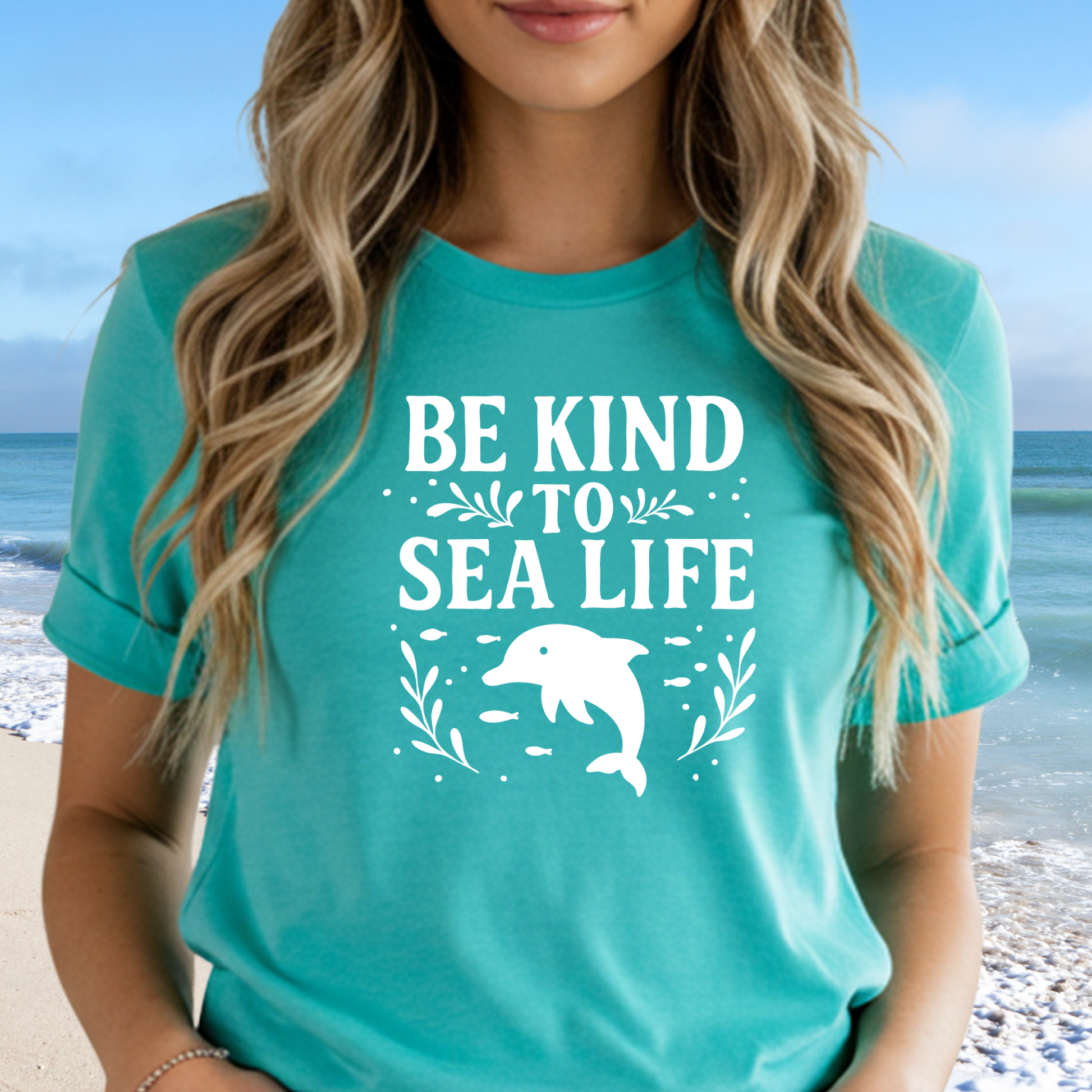 Person wearing a teal t-shirt with 'Be Kind to Sea Life' and a dolphin graphic on a beach.
