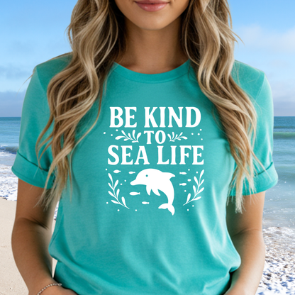 Person wearing a teal t-shirt with 'Be Kind to Sea Life' and a dolphin graphic on a beach.