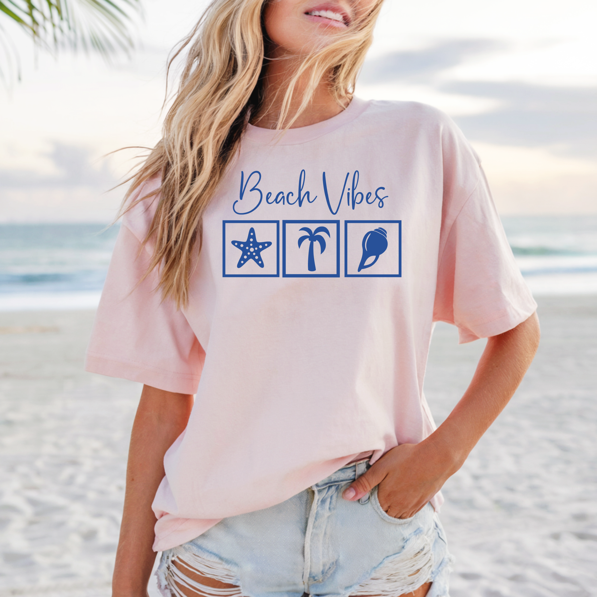 Bella and Canvas 3001 "Beach Vibes" t shirt with a starfish, palm tree, and a seashell graphics in monochrome color. Picture of a girl standing on the beach wearing a t shirt.