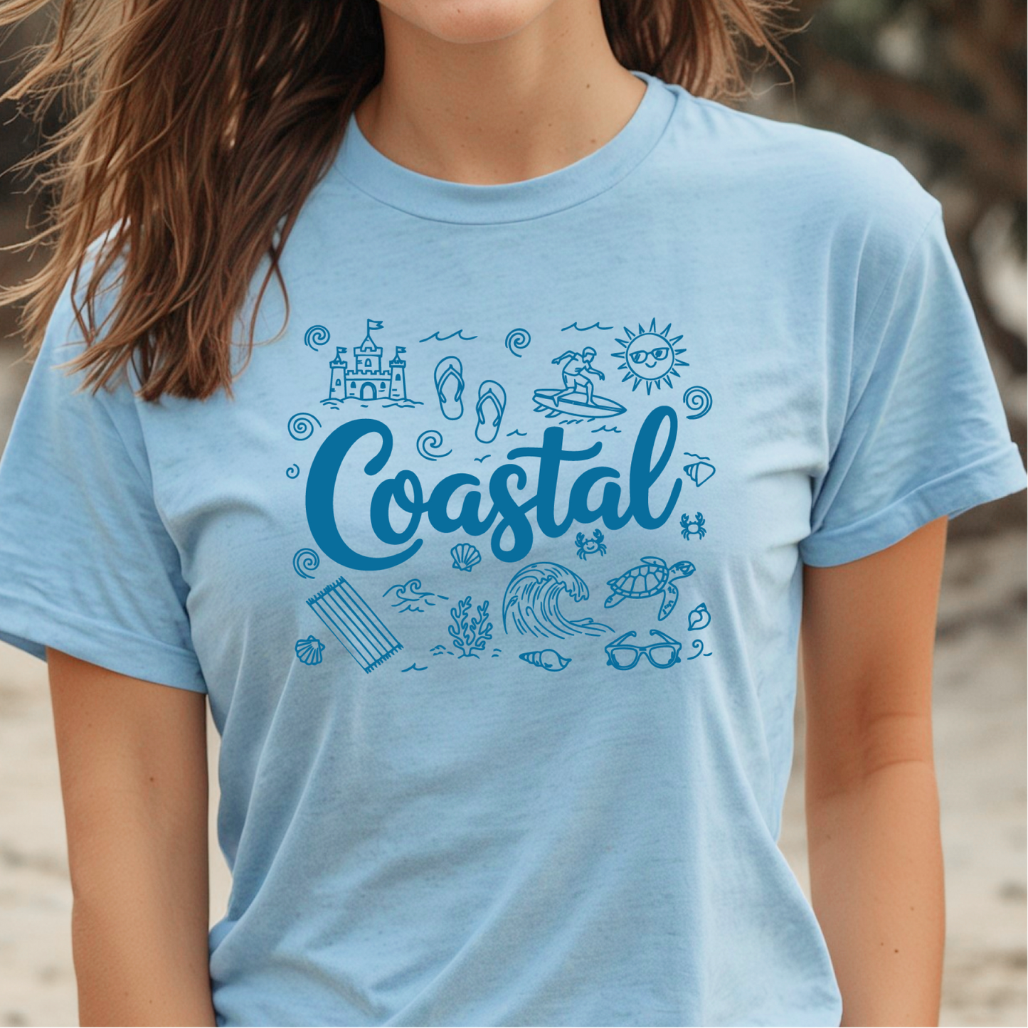 Person wearing a light blue t-shirt with 'Coastal' design on a beach.