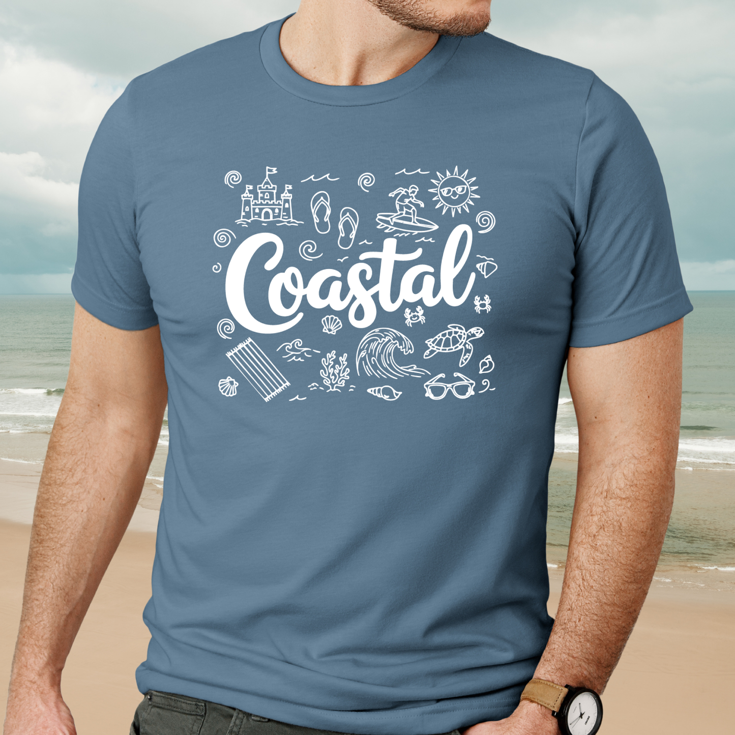 Person wearing a steel blue t-shirt with 'Coastal' design on a beach background
