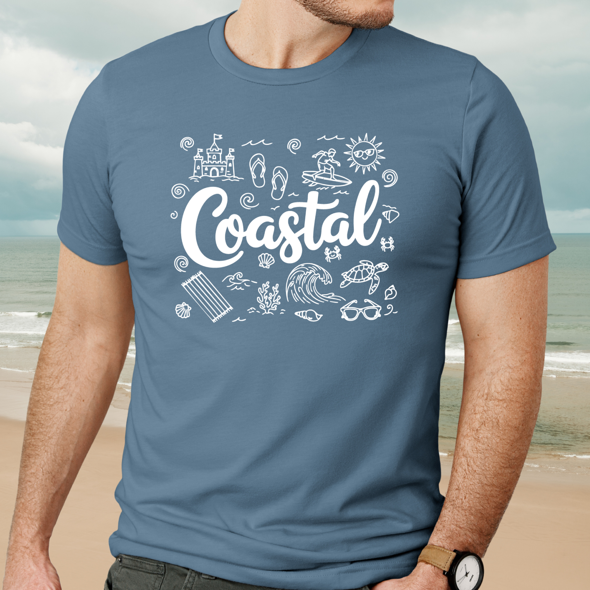 Person wearing a steel blue t-shirt with 'Coastal' design on a beach background