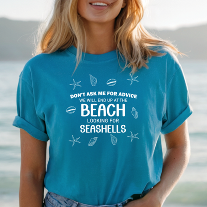 Person wearing a aqua blue t-shirt with beach-themed text and graphics, standing by a body of water.