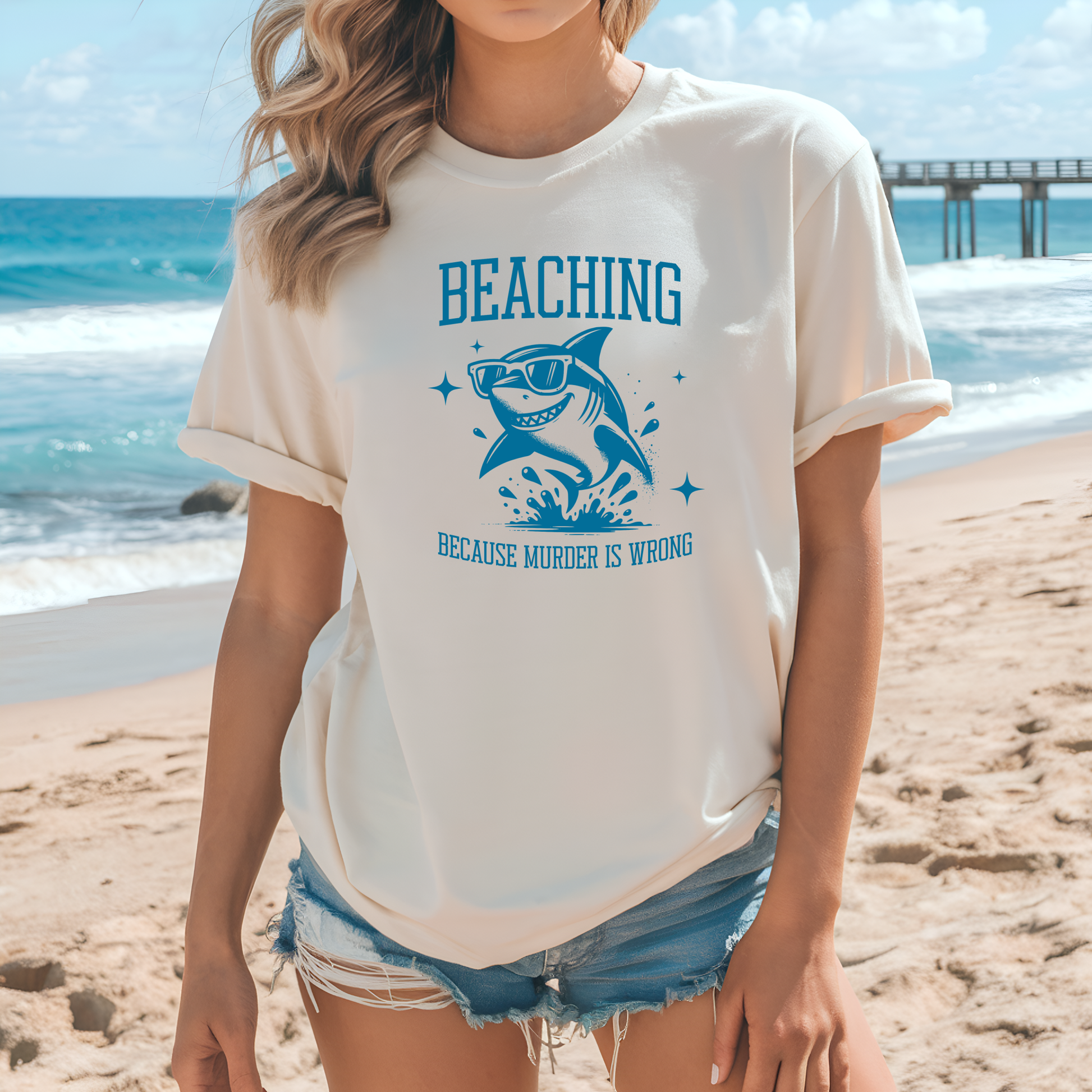 Bella Canvas 3001 Beaching Because Murder Is Wrong  T Shirt with Funny Shark with Sunglasses splashing in water in one color graphic. Picture of a girl standing on the beach wearing a t shirt.