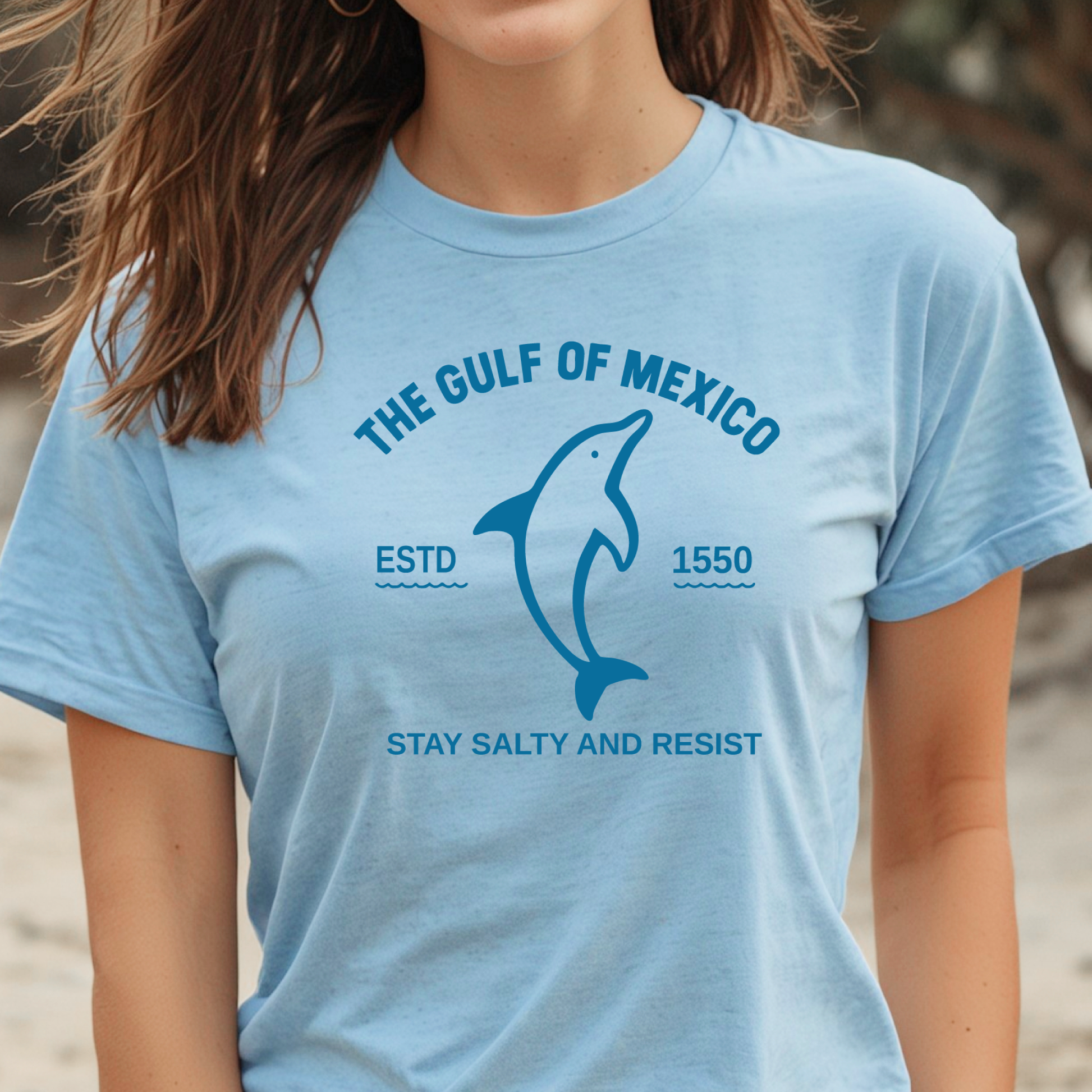 Person wearing a light blue t-shirt with a dolphin graphic and text on a beach.