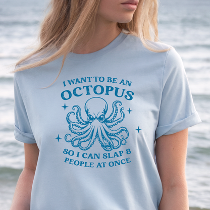 Person wearing a light blue t-shirt with an octopus graphic and text by a body of water.