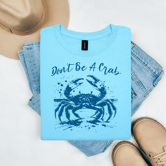 Gildan 5000 "Don't Be A Crab" t shirt with a crab standing on the beach in a one color graphic. Picture on a flay lay mockup with pair of blue jeans, brown shoes and a hat accessory.