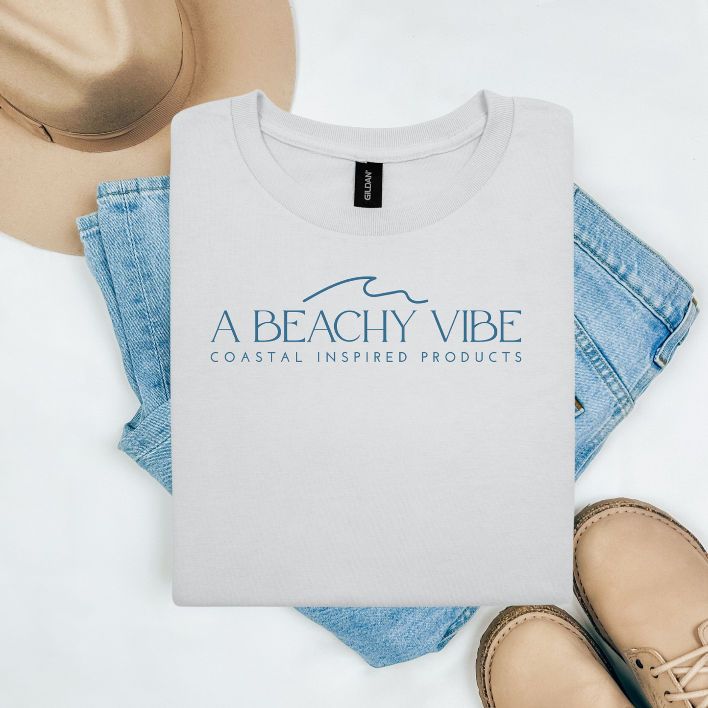 Gildan 5000 "A Beachy Vibe" t shirt with a simple wave graphic in one color. Picture on a flay lay mockup with pair of blue jeans, brown shoes and a hat accessory.