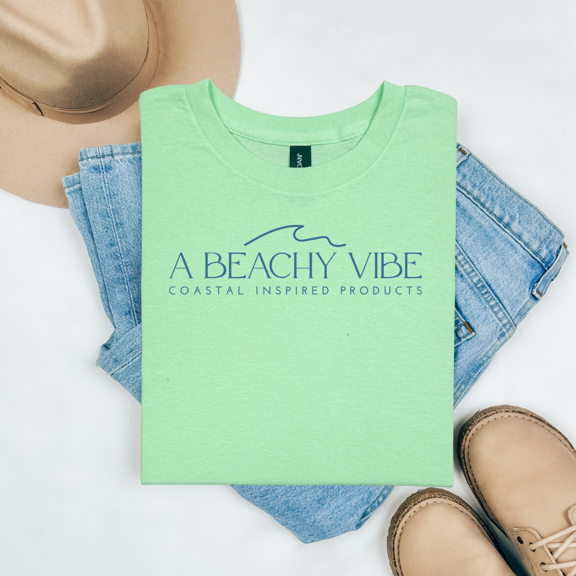 Gildan 5000 "A Beachy Vibe" t shirt with a simple wave graphic in one color. Picture on a flay lay mockup with pair of blue jeans, brown shoes and a hat accessory.