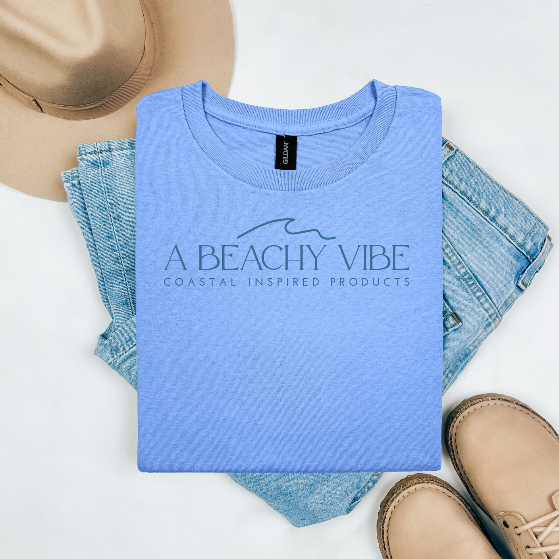 Gildan 5000 "A Beachy Vibe" t shirt with a simple wave graphic in one color. Picture on a flay lay mockup with pair of blue jeans, brown shoes and a hat accessory.