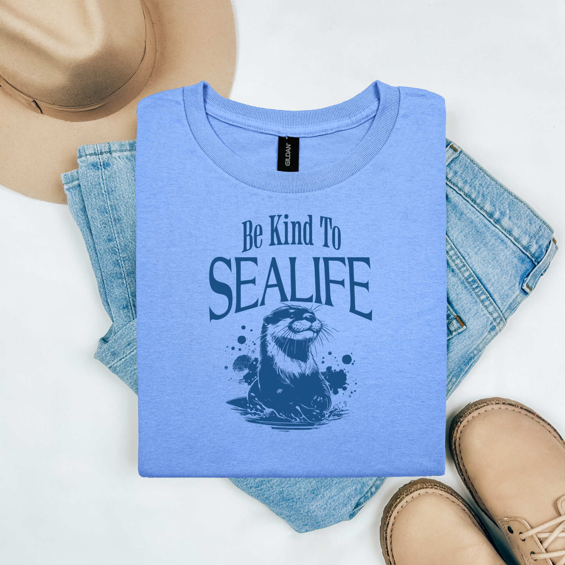 Gildan 5000 "Be Kind To Sealife" t shirt with a otter splashing in the water in a one color graphic. Picture on a flay lay mockup with pair of blue jeans, brown shoes and a hat accessory.