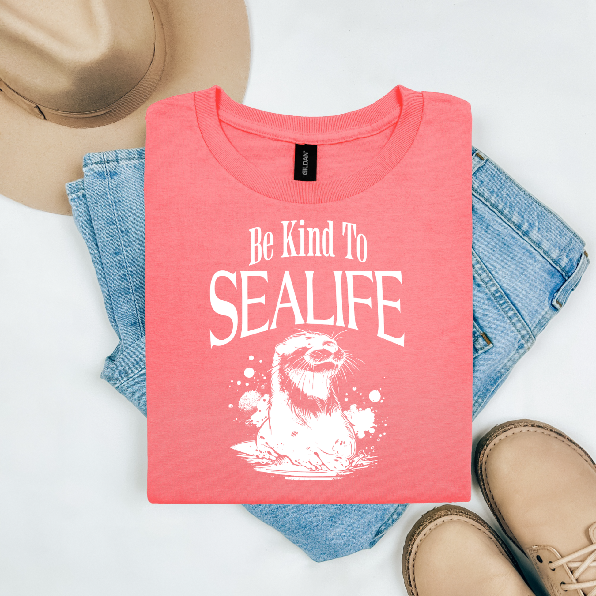 Gildan 5000 "Be Kind To Sealife" t shirt with a otter splashing in the water in a one color graphic. Picture on a flay lay mockup with pair of blue jeans, brown shoes and a hat accessory.