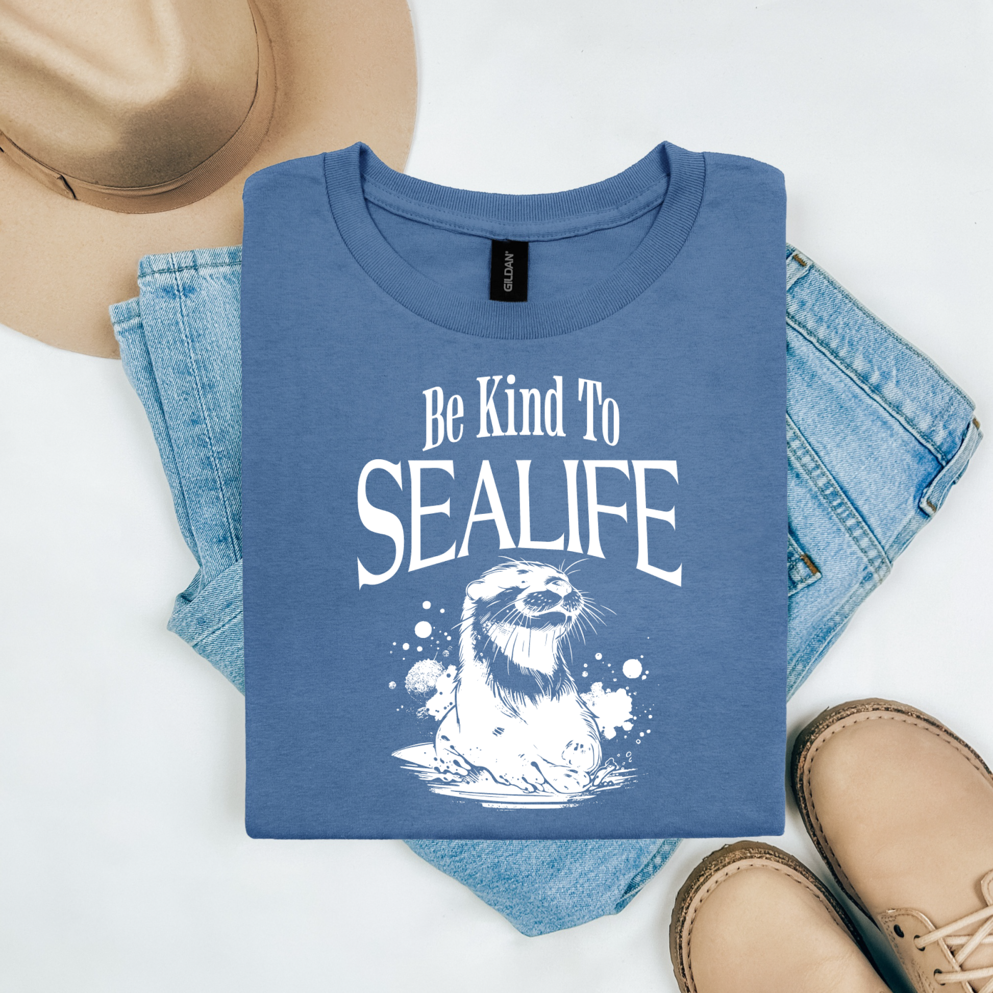 Gildan 5000 "Be Kind To Sealife" t shirt with a otter splashing in the water in a one color graphic. Picture on a flay lay mockup with pair of blue jeans, brown shoes and a hat accessory.