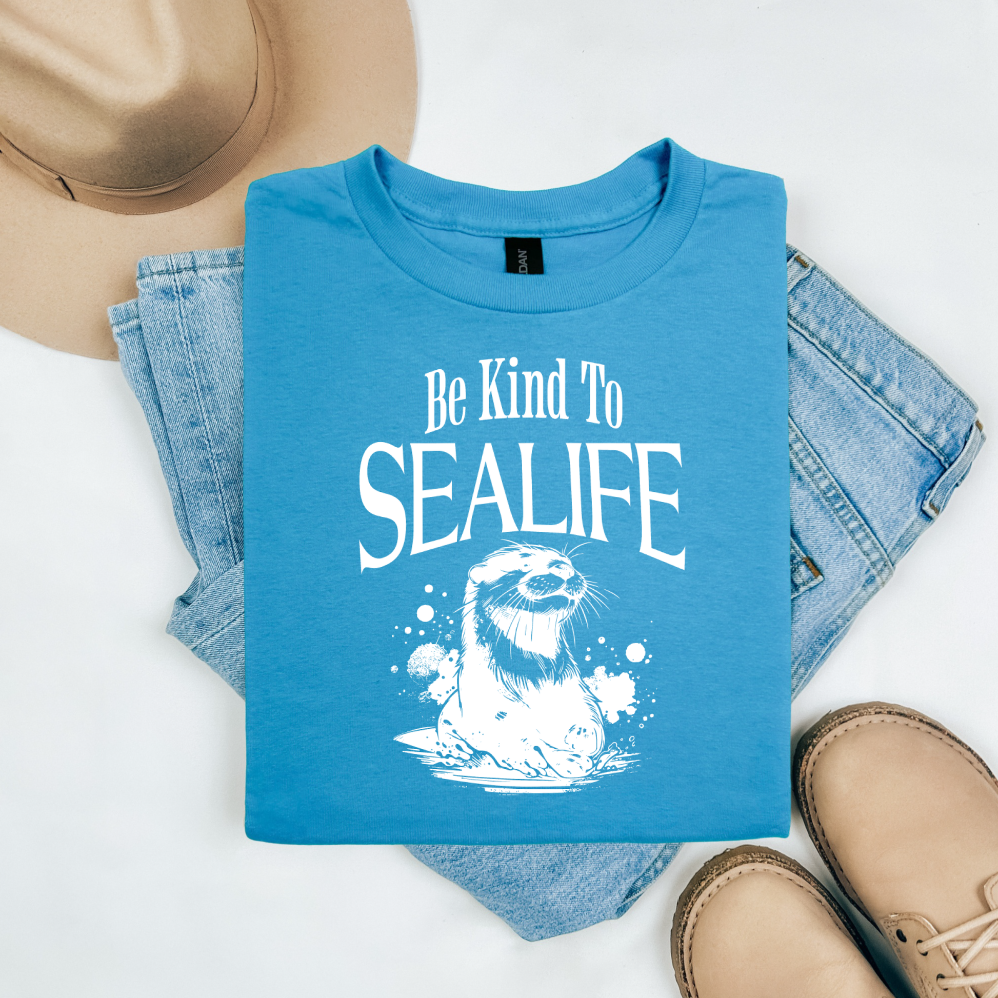 Gildan 5000 "Be Kind To Sealife" t shirt with a otter splashing in the water in a one color graphic. Picture on a flay lay mockup with pair of blue jeans, brown shoes and a hat accessory.