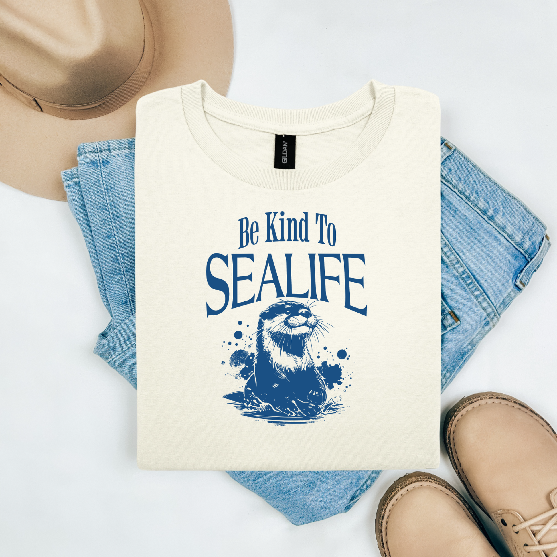 Gildan 5000 "Be Kind To Sealife" t shirt with a otter splashing in the water in a one color graphic. Picture on a flay lay mockup with pair of blue jeans, brown shoes and a hat accessory.