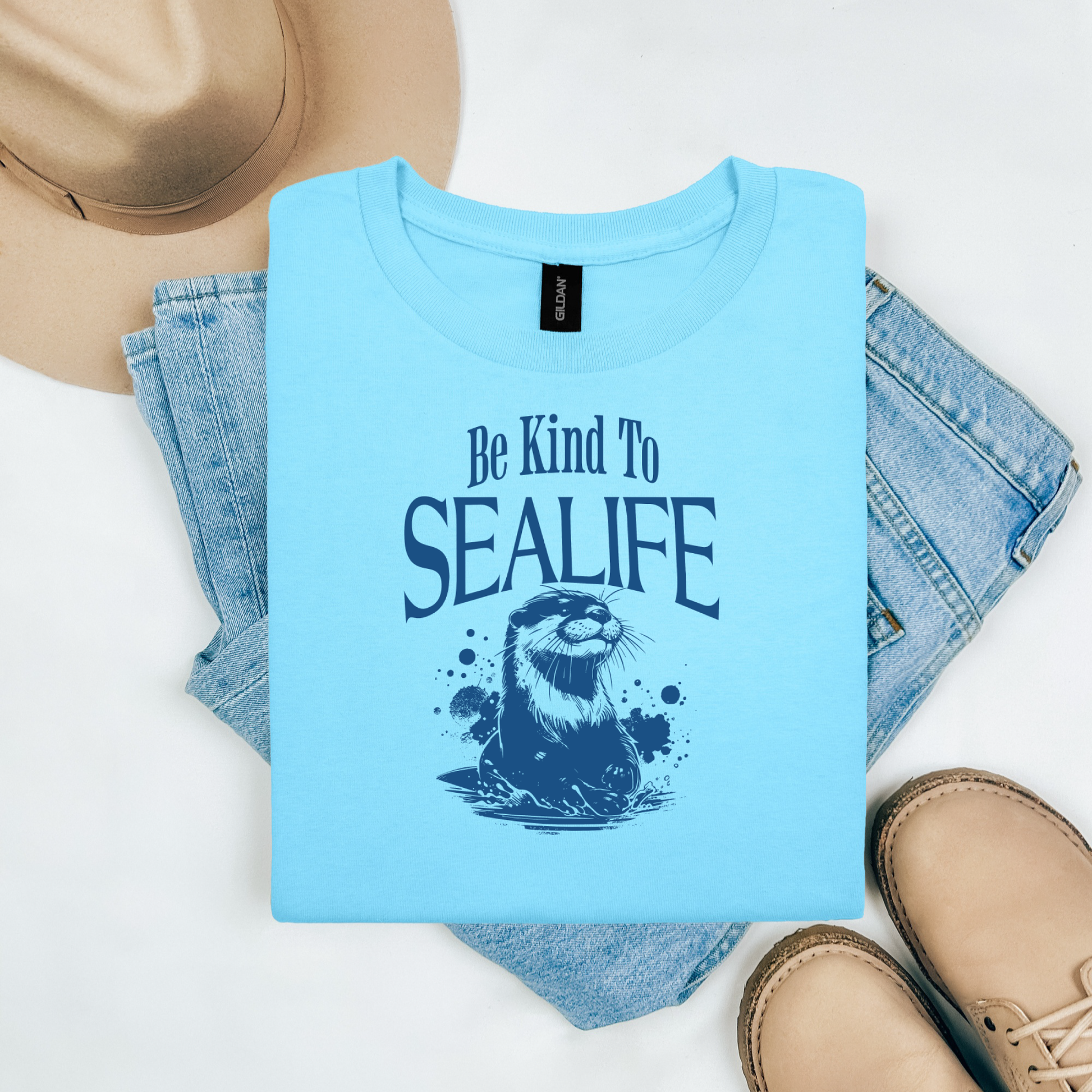 Gildan 5000 "Be Kind To Sealife" t shirt with a otter splashing in the water in a one color graphic. Picture on a flay lay mockup with pair of blue jeans, brown shoes and a hat accessory.