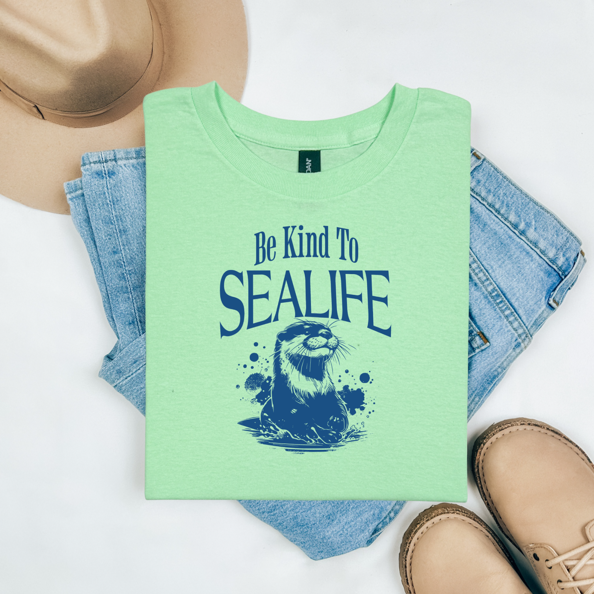 Gildan 5000 "Be Kind To Sealife" t shirt with a otter splashing in the water in a one color graphic. Picture on a flay lay mockup with pair of blue jeans, brown shoes and a hat accessory.