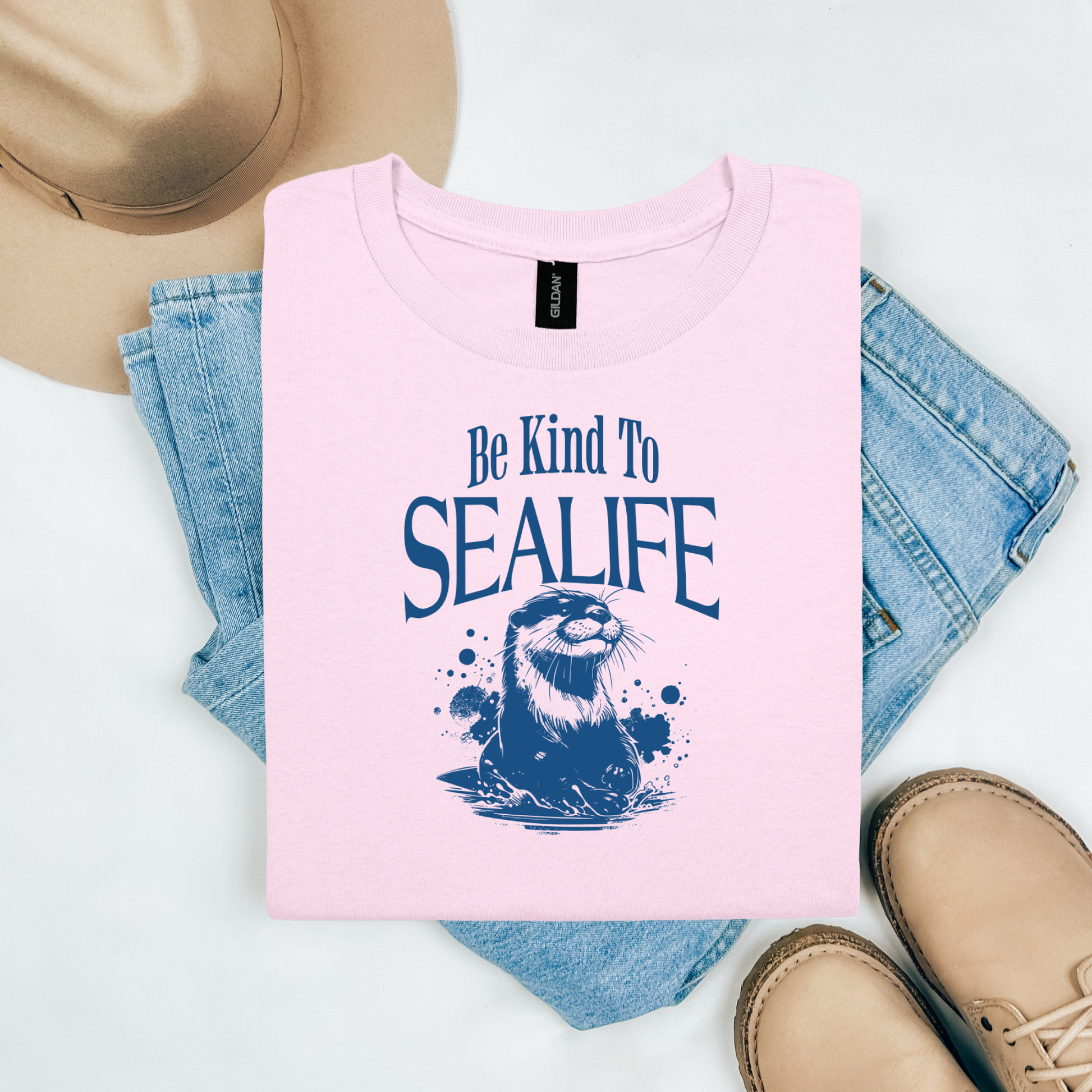 Gildan 5000 "Be Kind To Sealife" t shirt with a otter splashing in the water in a one color graphic. Picture on a flay lay mockup with pair of blue jeans, brown shoes and a hat accessory.