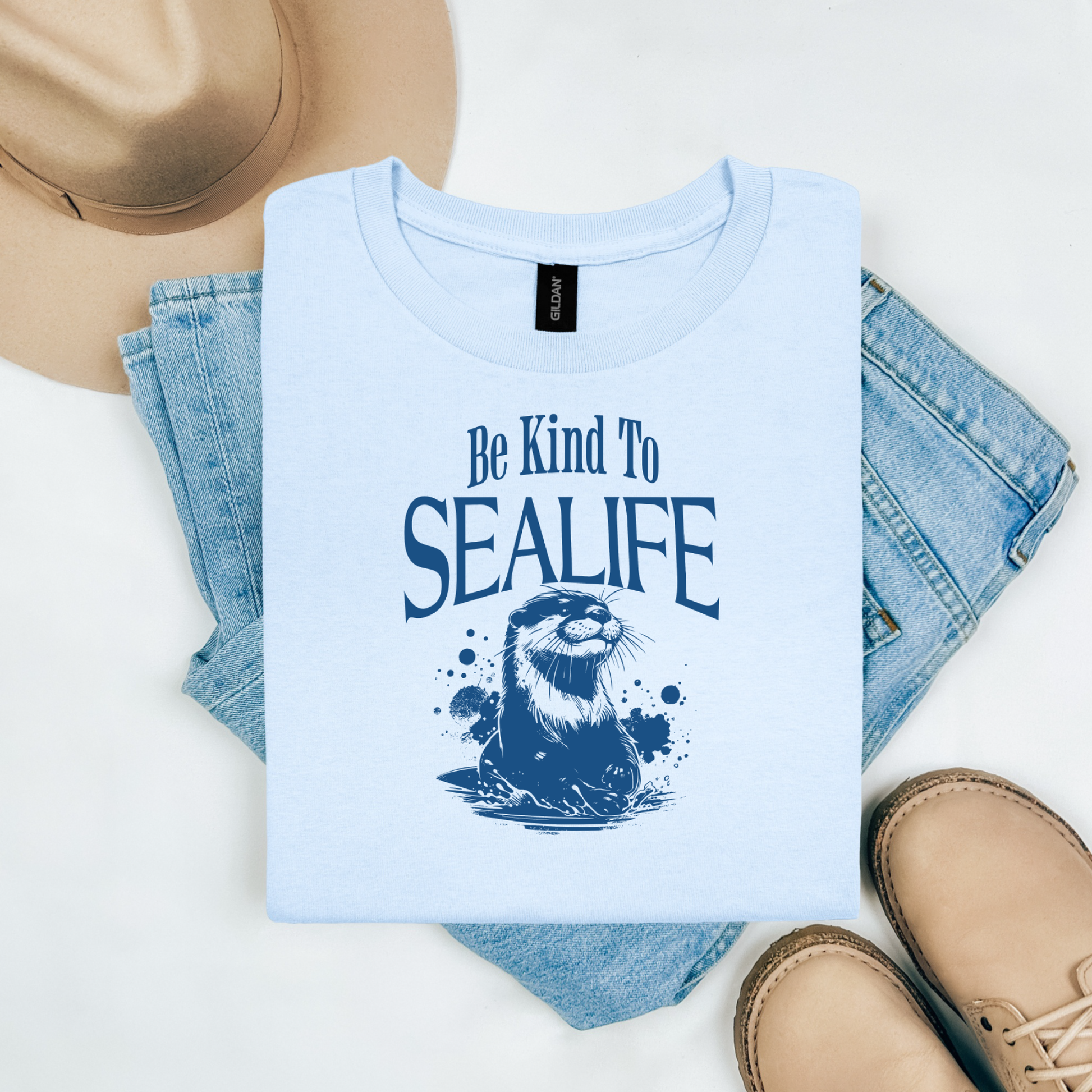 Gildan 5000 "Be Kind To Sealife" t shirt with a otter splashing in the water in a one color graphic. Picture on a flay lay mockup with pair of blue jeans, brown shoes and a hat accessory.